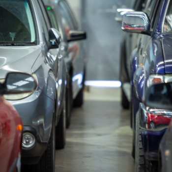 A narrow corridor between two rows of cars parked closely together inside an automotive workshop, with soft light visible in the background
